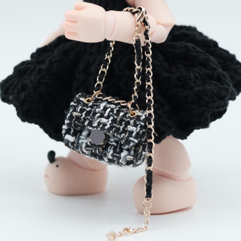 May include: A miniature black and white tweed handbag with a gold chain strap. The small purse has a black flower-shaped clasp and is held by a doll. The doll is wearing a black dress.