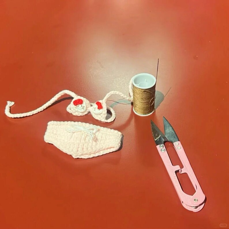 Puede incluir: Un conjunto de bikini de ganchillo hecho a mano en rosa claro y blanco, con detalles florales rojos. Una bobina de hilo dorado, una aguja y unas tijeras rosas tambi&eacute;n est&aacute;n presentes sobre una superficie roja. El top y la braguita del bikini se muestran por separado.