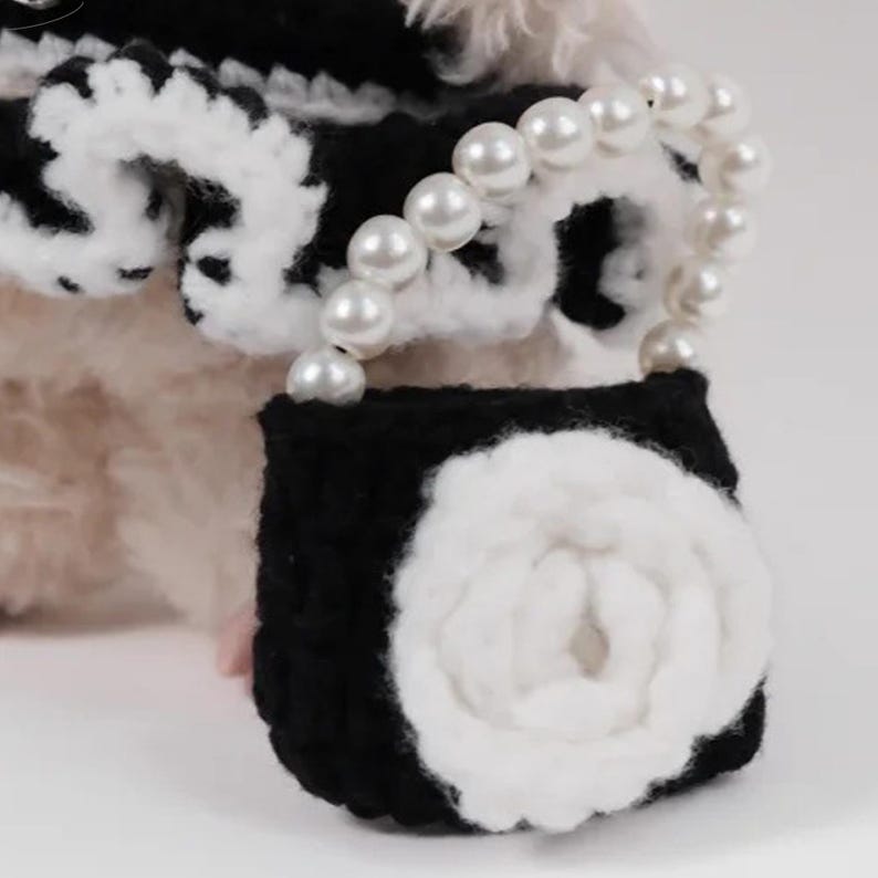 May include: A black and white crocheted purse with a white flower design and a pearl handle. The purse is small and has a decorative, feminine style. The background is a soft, neutral color.