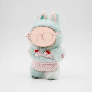 Puede incluir: Mu&ntilde;eca de peluche con un disfraz de conejito azul claro con detalles en rosa. La mu&ntilde;eca lleva gafas redondas rosas y un conjunto de ganchillo rosa. El disfraz de conejito tiene orejas largas y una capucha.