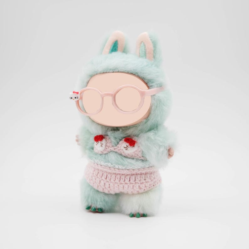 Puede incluir: Mu&ntilde;eca de peluche con un disfraz de conejito azul claro con detalles en rosa. La mu&ntilde;eca lleva gafas redondas rosas y un conjunto de ganchillo rosa. El disfraz de conejito tiene orejas largas y una capucha.