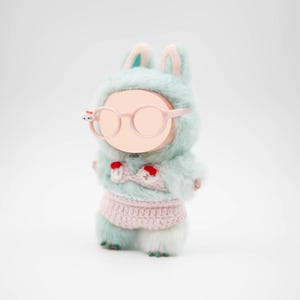 Puede incluir: Una figura de conejo de peluche con un atuendo con capucha azul claro y gafas rosas. La figura tiene una parte superior e inferior de ganchillo rosa con detalles en rojo. Los pies del conejo son verdes.