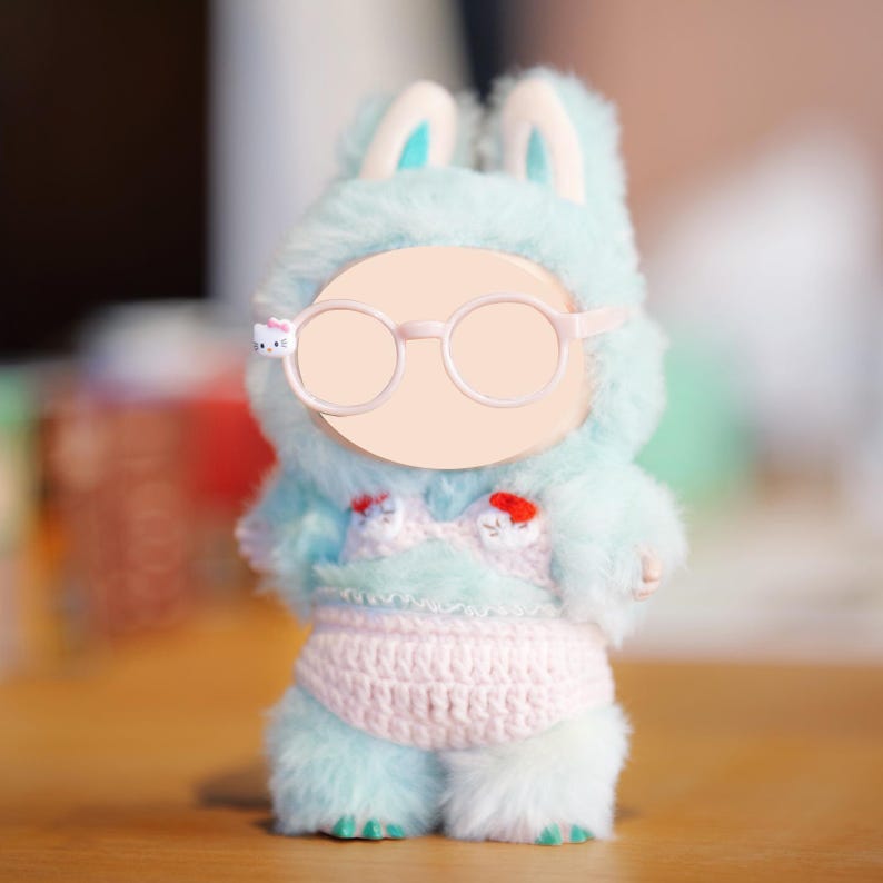 Puede incluir: Una figura de conejito de peluche con un traje de piel azul claro con orejas de conejo a juego. La figura lleva un top de bikini y un pa&ntilde;al de ganchillo rosa. Tiene una cara en blanco con gafas redondas y un detalle de Hello Kitty en la montura.