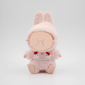 Puede incluir: Una mu&ntilde;eca de peluche rosa con un gorro de conejo a juego, gafas y un conjunto de ganchillo. La mu&ntilde;eca tiene una cara rosa p&aacute;lido y peque&ntilde;os detalles rojos en el conjunto. Las gafas tienen un peque&ntilde;o detalle blanco y negro en el lateral.