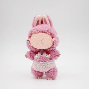 Puede incluir: Una mu&ntilde;eca de peluche rosa con capucha de orejas de conejo, gafas redondas y un atuendo de ganchillo. El atuendo de la mu&ntilde;eca incluye una braga de ganchillo blanca y una parte superior con detalles de coraz&oacute;n rojo. La mu&ntilde;eca est&aacute; sobre un fondo blanco.