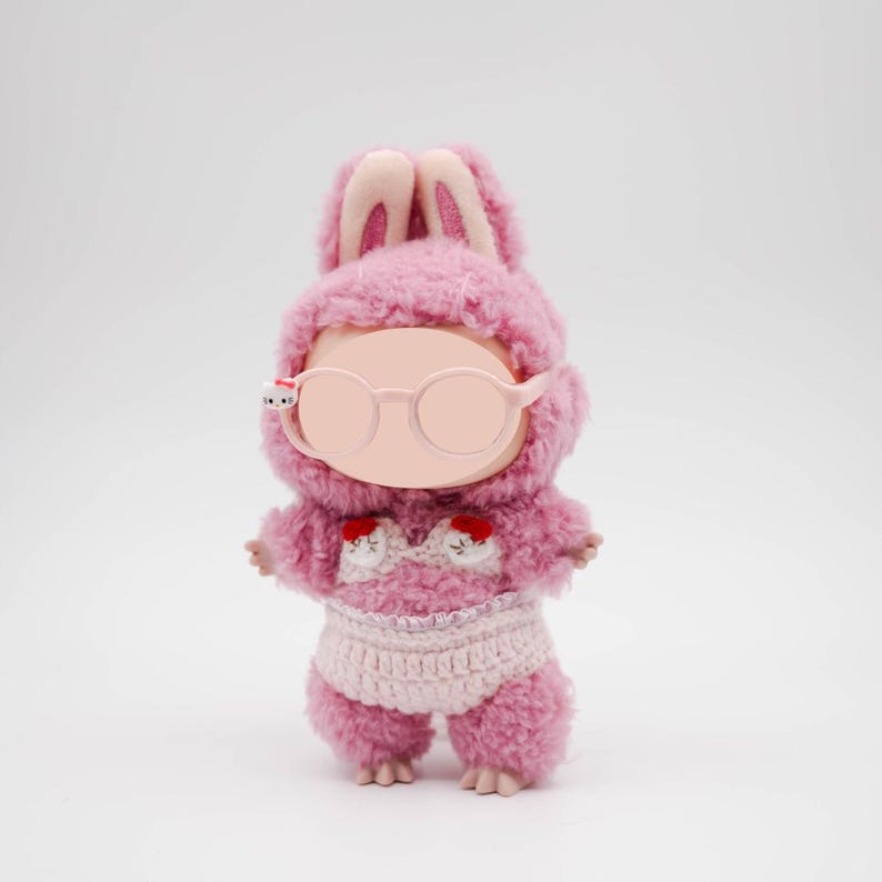 Puede incluir: Una mu&ntilde;eca de peluche rosa con capucha de orejas de conejo, gafas redondas y un atuendo de ganchillo. El atuendo de la mu&ntilde;eca incluye una braga de ganchillo blanca y una parte superior con detalles de coraz&oacute;n rojo. La mu&ntilde;eca est&aacute; sobre un fondo blanco.