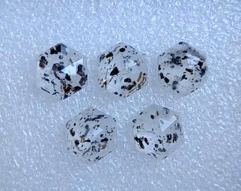 May include: Five clear, hexagonal gemstones with black and brown inclusions are arranged in a box. The gems have multiple facets, reflecting light. The gems are set against a white, textured background.