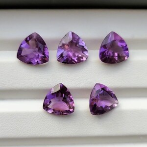 May include: Five triangular amethyst gemstones. The gemstones are a deep purple color with lighter purple and pink hues. The stones are faceted and have a polished surface. They are arranged on a white surface.