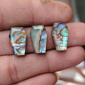 May include: Three iridescent coffin-shaped gemstones with faceted surfaces. The gemstones display a mix of colors, including blues, greens, and browns, with a pearlescent sheen. The gemstones are held in a hand.