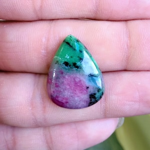 Puede incluir: Una gema pulida en forma de lágrima. La piedra presenta una mezcla de colores verdes, magenta y negros. La gema se sostiene en una mano, mostrando su forma y tamaño. Es probable que sea un cabujón.