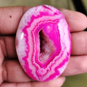 May include: Oval-shaped polished agate gemstone with a vibrant pink and white banded pattern. The center features a geode cavity with sparkling crystals. The stone is held in a hand, showcasing its size and natural beauty.