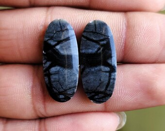 May include: Two oval-shaped gemstones with a dark gray and black pattern. The stones have a polished surface and are held in a hand. The pattern resembles a network of black veins against a gray background. The gemstones are likely for jewelry making.