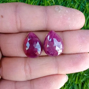 May include: Two faceted, teardrop-shaped rubies. The gems are a deep, rich red color and have multiple facets that catch the light. The rubies are displayed against a background of a hand and green grass.