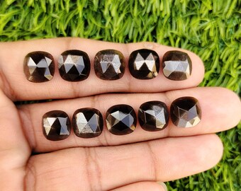 Pode incluir: Dez pedras preciosas de corte quadrado, castanho escuro, com superfícies facetadas, são dispostas sobre uma mão sobre um fundo verde. As pedras têm uma qualidade reflexiva, captando a luz e exibindo vários padrões internos.