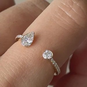 May include: Gold-toned open ring featuring a pear-cut diamond and a round diamond. The band is embellished with small accent stones. The diamonds are clear and reflect light.