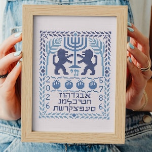 May include: A framed cross-stitch featuring a blue and white design. The artwork includes a menorah, lions, and Hebrew text. The frame is made of light-colored wood. The piece is held by a person wearing a denim overall.