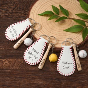 May include: Three baseball-themed keychains featuring white leather teardrop shapes with red stitching, small wooden bats engraved with names, and miniature baseballs or a yellow softball attached by silver metal rings and chains.