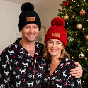 Puede incluir: Una pareja con gorros de punto negros y rojos a juego con pompones de piel sintética y etiquetas con nombres. También visten pijamas negros a juego con un estampado de renos y copos de nieve blancos. Un árbol de Navidad está al fondo.