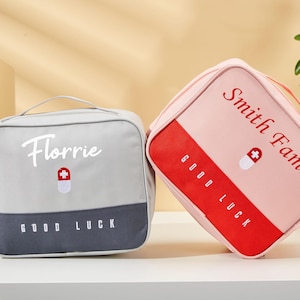 May include: Two square first aid kits. One is gray with the name "Florrie" and a pill icon, the other is pink with "Smith Family" and a pill icon. Both have "GOOD LUCK" printed on them.