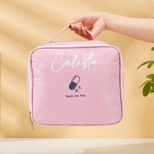 Könnte beinhalten: Eine rosa Medizintasche mit Griff. Auf der Tasche steht der Name "Calista" in Schreibschrift, eine Pillengrafik und die Worte "Medicine bag". Unten ist ein Reißverschluss zu sehen. Die Tasche wird von einer Person gehalten.