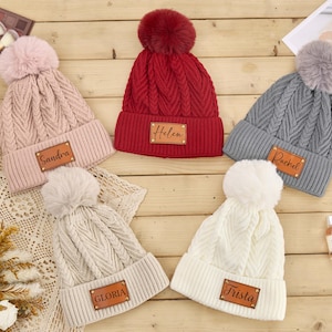 May include: Five cable-knit beanies in pink, red, gray, white, and cream, each with a faux fur pom-pom. The beanies have a leatherette patch with a name on each: Sandra, Helen, Rachel, Gloria, and Trista. The hats are on a wooden surface.