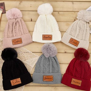 Peut inclure: Six bonnets en tricot avec des pompons en fausse fourrure. Chaque bonnet est de couleur différente : rose, blanc, beige, noir, gris et rouge. Chaque bonnet porte une étiquette rectangulaire en cuir avec un nom. Présentés sur une surface en bois.