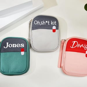 May include: Three small, zippered pouches in teal, grey, and pink. Each pouch has a two-tone design with a contrasting top section and a white pill graphic. The pouches are labelled with the words "Jones", "Oh, sh*t kit", and "Doris".