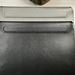 Puede incluir: Dos fundas de cuero para portátiles, una gris y otra negra, apiladas. Cada funda tiene un cierre de solapa con borde redondeado. Las fundas están sobre una superficie de madera clara.