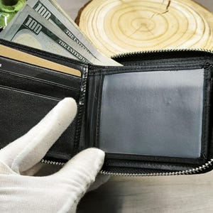May include: Black leather wallet with a zip closure, open to show a clear ID window, card slots, and cash. The wallet contains several one hundred dollar bills and a gold-coloured card. The word "BLACK" is visible on a card slot.