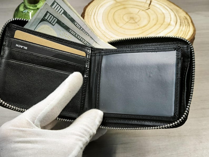 May include: Black leather wallet with a zip closure, open to show a clear ID window, card slots, and cash. The wallet contains several one hundred dollar bills and a gold-coloured card. The word "BLACK" is visible on a card slot.