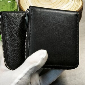 May include: Black leather wallet with a zippered compartment and a smooth, rectangular design. The wallet is held by a gloved hand, showcasing its sleek and minimalist aesthetic. The wallet is a practical accessory for carrying cards and cash.