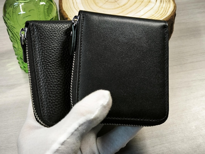 May include: Black leather wallet with a zippered compartment and a smooth, rectangular design. The wallet is held by a gloved hand, showcasing its sleek and minimalist aesthetic. The wallet is a practical accessory for carrying cards and cash.