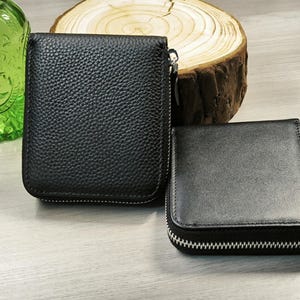 May include: Two black leather wallets with zip closures. One wallet is rectangular and larger, while the other is square and smaller. The wallets are displayed on a light-coloured surface, with a green glass bottle and a wood slice in the background.