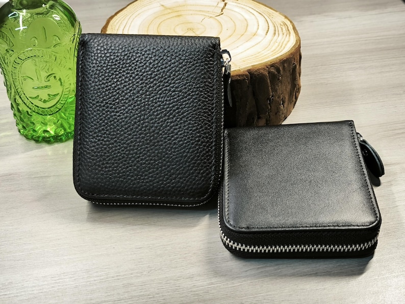 May include: Two black leather wallets with zip closures. One wallet is rectangular and larger, while the other is square and smaller. The wallets are displayed on a light-coloured surface, with a green glass bottle and a wood slice in the background.