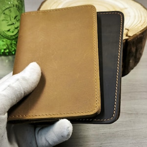 May include: Two leather wallets, one tan and one dark brown, are stacked. The tan wallet is in the foreground, with visible stitching around the edges. The wallets are held by a gloved hand. A green glass and a wood slice are in the background.