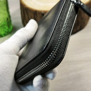 May include: Black leather wallet with a silver zip. The wallet is rectangular and appears to be closed. The zip runs along three sides of the wallet. The wallet is held by a gloved hand.