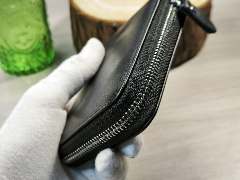 May include: Black leather wallet with a silver zip. The wallet is rectangular and appears to be closed. The zip runs along three sides of the wallet. The wallet is held by a gloved hand.