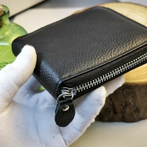May include: A black, square, zippered wallet with a textured leather exterior. The silver zip runs along three sides. The wallet is held by a gloved hand. A green glass bottle with a cork stopper is in the background.
