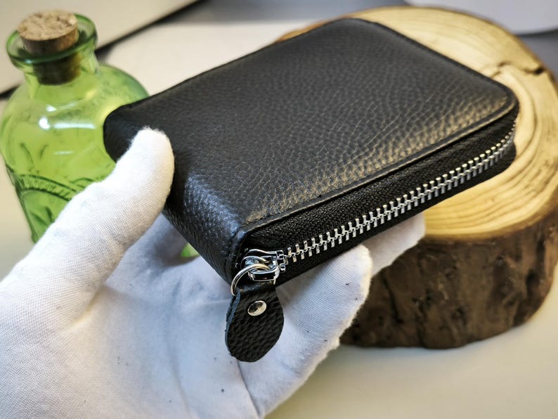 May include: A black, square, zippered wallet with a textured leather exterior. The silver zip runs along three sides. The wallet is held by a gloved hand. A green glass bottle with a cork stopper is in the background.