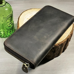 May include: A dark brown leather wallet with a zipper closure. The wallet is rectangular and appears to be made of distressed leather. The wallet is approximately 8 inches long and 4 inches wide.