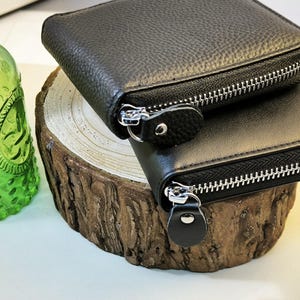 May include: Two black leather wallets with silver zips are stacked on a wooden surface. The top wallet is smaller than the bottom one. A green glass bottle with a decorative pattern is visible in the background.