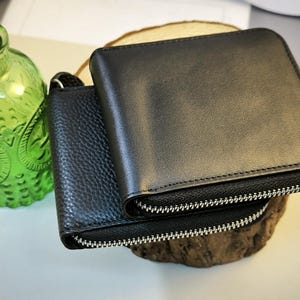 May include: Two black leather wallets with silver zips are stacked on a wooden slice. The top wallet is smooth, while the bottom one has a textured surface. A green glass bottle with a cork stopper is in the background.