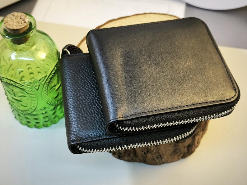 May include: Two black leather wallets with silver zips are stacked on a wooden slice. The top wallet is smooth, while the bottom one has a textured surface. A green glass bottle with a cork stopper is in the background.