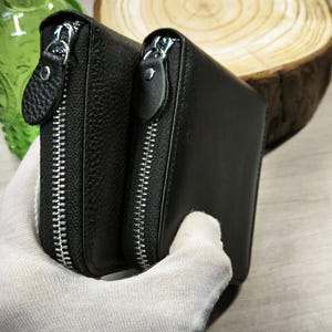 May include: Two black leather wallets with silver zips. The wallets have a textured surface and are held together. The background includes a green glass object and a wooden slice.
