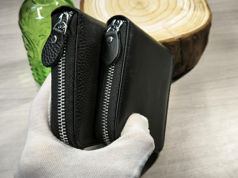 May include: Two black leather wallets with silver zips. The wallets have a textured surface and are held together. The background includes a green glass object and a wooden slice.