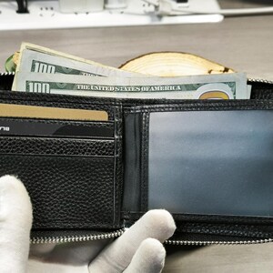 May include: Open black leather wallet with a zip closure, containing several one hundred dollar bills and a credit card. The wallet has multiple compartments and a clear ID window. The word "BLACK" is visible on the credit card.