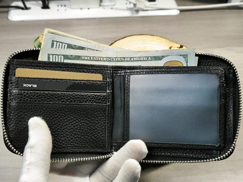 May include: Open black leather wallet with a zip closure, containing several one hundred dollar bills and a credit card. The wallet has multiple compartments and a clear ID window. The word "BLACK" is visible on the credit card.