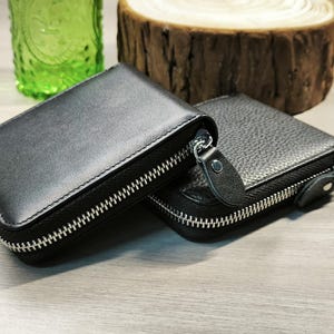 May include: Two black leather wallets with silver zips. One wallet has a smooth texture, while the other has a pebbled texture. The wallets are rectangular and appear to be closed. The background includes a green glass and a wooden slice.