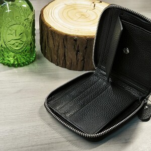 May include: Black leather zip-around wallet, open to show card slots and compartments. The wallet has a silver zip and a small pull tab. A green glass bottle and a wood slice are in the background.