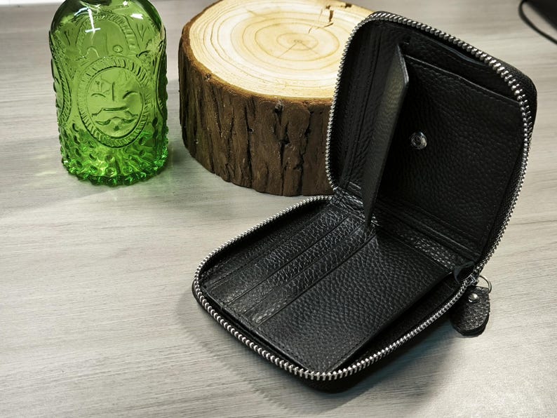 May include: Black leather zip-around wallet, open to show card slots and compartments. The wallet has a silver zip and a small pull tab. A green glass bottle and a wood slice are in the background.